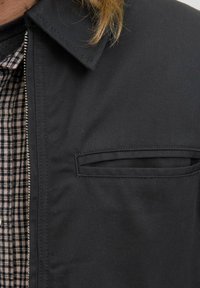 Gros plan sur le col d'une veste noire et la poche de poitrine sur une chemise à carreaux, avec de longs cheveux blonds visibles près du col.