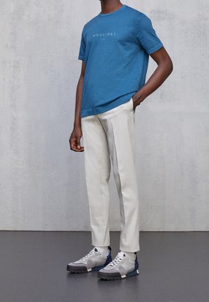 Homme portant un t-shirt bleu à manches courtes, un pantalon écru et des baskets grises et blanches, debout devant un fond uni.