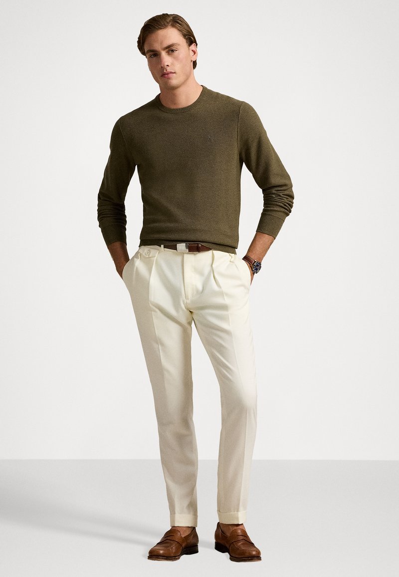 Maglione a maglia verde oliva, pantaloni bianchi plissettati, scarpe in pelle marrone e un orologio con cinturino marrone, esposti su uno sfondo neutro.