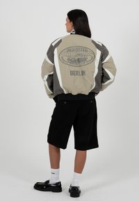 Personne aux longs cheveux foncés portant une veste de course beige et grise avec le texte "Prohibited Racing Club Berlin", un short noir, des chaussettes blanches et des chaussures noires.