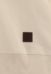 Tejido beige con una textura suave presenta un parche rectangular de cuero marrón etiquetado "KEY LARGO". Diseño simple y minimalista.