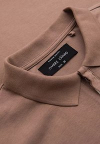 Camisa polo marrón con cuello acanalado, que presenta un pequeño cierre con cremallera y una etiqueta de cuidado que dice "ombre eximmo, talla M." Textura suave.