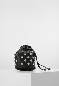 Borsa a secchio nera in pelle con borchie argentate, raccolta nella parte superiore, posizionata su un piedistallo bianco contro uno sfondo semplice.