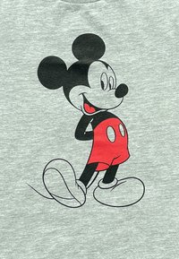 Grijze T-shirt met een zwart en rood cartoonfiguur met ronde oren, die glimlacht en poseert, tegen een gestructureerde achtergrond.