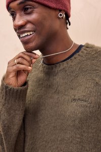 Olivengrøn strikket sweater med en tekstureret overflade, rund halsudskæring og broderet logo. Sølvkæde omkring halsen, rød strikket hue.