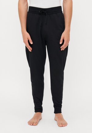 Schwarze Jogginghosen mit einem Kordelzugbund, tailliertem Schnitt und weicher Textur. Keine sichtbaren Muster oder Logos. Gestylt mit nackten Füßen.