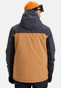Mann in einer zweifarbenen braunen und dunkelgrauen Winterjacke, trägt eine leuchtend orangefarbene Mütze und schwarze Skibrille auf dem Kopf, wendet sich ab.