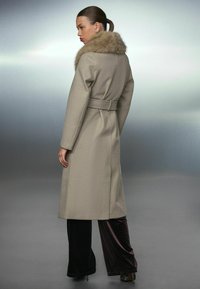 Beige wollen blend jas met een bontkraag, een ceintuur in de taille en lange mouwen, gecombineerd met donkerbruine satijnen broeken, gepresenteerd vanuit een achterkant perspectief.