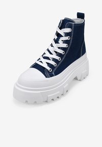 Zapatillas de denim azul de caña alta con suelas de goma blanca, que cuentan con una plataforma gruesa y punta redonda. Atadas con cordones blancos y con ojetes reforzados.