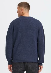 Pull en maille marine avec un motif texturé, manches longues et encolure côtelée, vu de dos sur un fond clair.