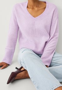 Maglione viola chiaro a scollo a V in tessuto lavorato a maglia con polsini e orlo a coste, indossato con jeans in denim blu e scarpe a punta di colore rosso scuro.