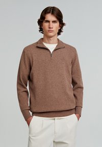 Brun strikket sweatshirt med lynlås og høj hals, lange ærmer, ribbede manchetter og kant, båret over en hvid skjorte, parret med lyse bukser.