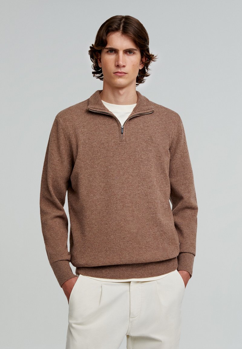 Brun strikket sweatshirt med lynlås og høj hals, lange ærmer, ribbede manchetter og kant, båret over en hvid skjorte, parret med lyse bukser.