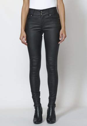 Kvinde iført sorte højtaljede skinny bukser i imiteret læder og sorte ankelstøvler, stående foran en ensfarvet baggrund.