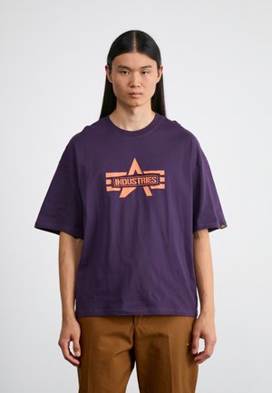 Jeune personne aux longs cheveux foncés, portant un T-shirt violet ample avec un logo orange "Industries" et un pantalon marron, debout devant un fond blanc.