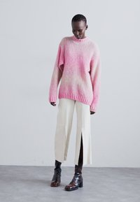 Maglione oversize con gradiente rosa e polsini a coste, abbinato a pantaloni larghi color crema con spacchi sui lati. Stivali neri lucidi alla caviglia completano l'outfit.