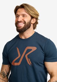 Mörkblå bomullströja med en orange randig logotyp. Korta ärmar, rund hals och en figurnära design med synliga tatueringar på armen.