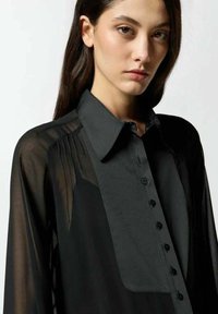 Camicia nera con bottoni, dotata di un pannello frontale solido e maniche trasparenti. Presenta un collare classico e pieghe verticali sulle spalle.