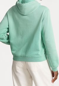 Mátově zelená mikina s kapucí, texturovanou tkaninou, žebrovanými manžetami a spodním lemem, a volným střihem, spárovaná se světlými kalhotami.