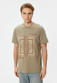 Olivenfarvet bomulds t-shirt med grafisk design med tribal mønstre og teksten "BE BRAVE". Korte ærmer og rund halsudskæring.