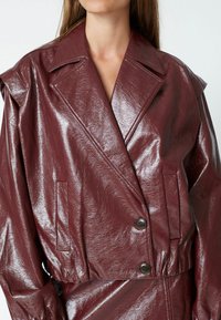 Giacca in finta pelle borgogna con finitura lucida, colletto oversize, due tasche frontali e chiusura a doppio bottone.