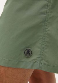 Groene zwembroek van lichtgewicht materiaal, met een zwarte ronde patch met een doodskop en gekruiste botten logo aan de onderzoom.