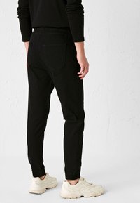 Pantaloni jogger neri con vita elastica, tasche posteriori e polsini elastici, abbinati a scarpe da ginnastica chiare su una superficie testurizzata.