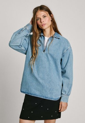 Lichtblauwe denim pullover sweatshirt met een halve rits, een kraag en lange mouwen. Gecombineerd met een zwarte rok met decoratieve studs.