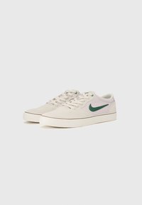 Tenisi cu superior din piele de-suede bej deschis, având un logo Nike swoosh verde, șireturi plate și o talpă din cauciuc alb.