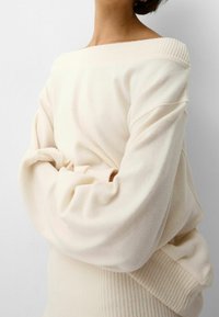 Kremowy, oversized sweter z prążkowanym dekoltem i mankietami, z miękką fakturą i luźnym krojem, który delikatnie opada na ramionach.