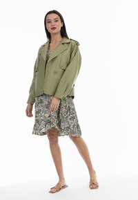 Giacca oversized in eco-pelle verde con bottoni doppi, indossata sopra un vestito floreale, abbinata a sandali beige. Sfondo bianco.