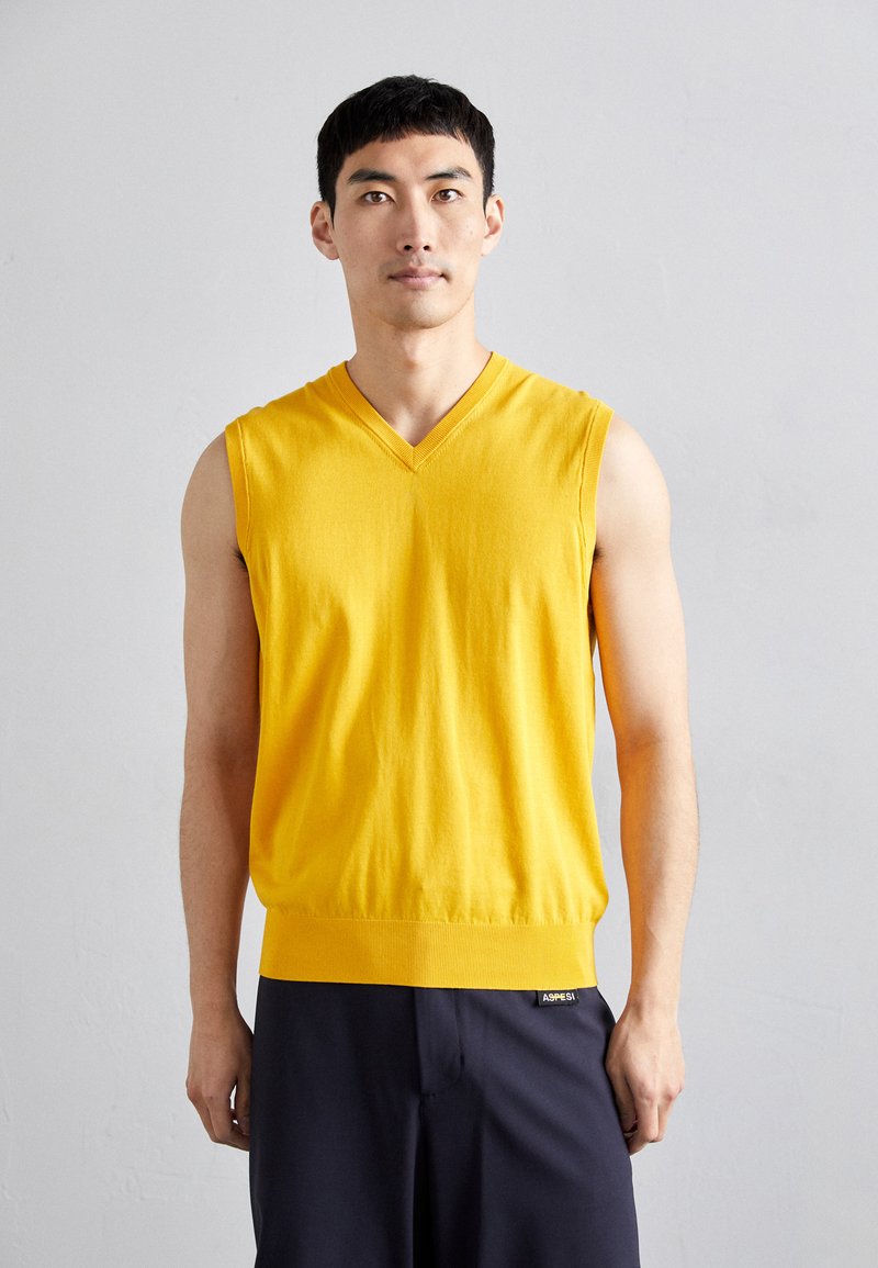 Yellow sleeveless sweater in soft knit fabric, featuring a v-neck design and ribbed bottom hem, worn over dark pants.