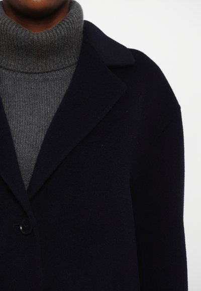 Manteau en laine bleu marine foncé avec un col, un seul bouton et un pull col roulé en gris doux à côtes en dessous. Texture lisse, coupe décontractée.