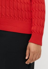 Rød strikket genser med en strukturert flette-design, ribbestrikkede mansjetter og kant. Stoffet ser mykt ut med en glatt overflate.