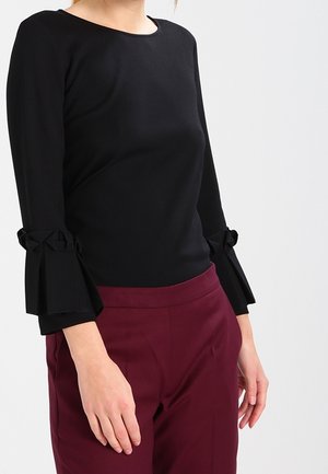 Kobieta ubrana w czarną bluzkę z długim rękawem z kokardkami na rękawach oraz bordowe spodnie z wysokim stanem, stojąca na jednolitym tle.