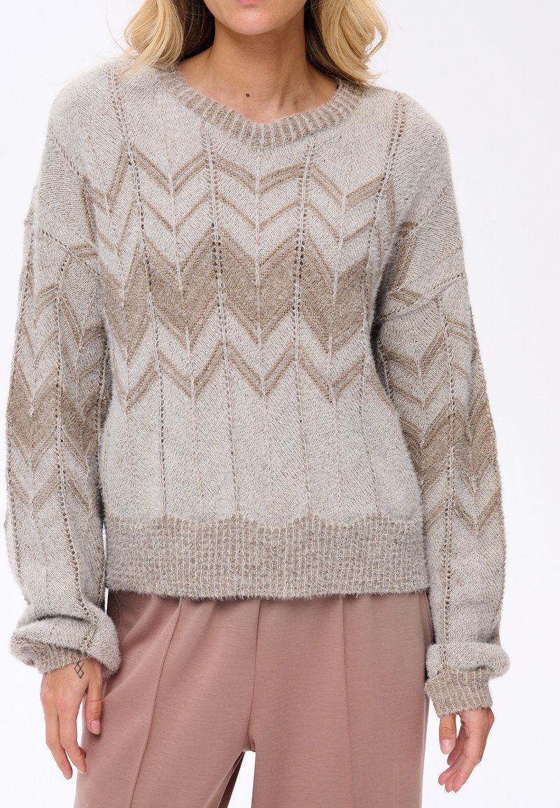 Vrouw in een beige en bruin gebreid vest met een zigzagpatroon en mauve broek, staand tegen een witte achtergrond.