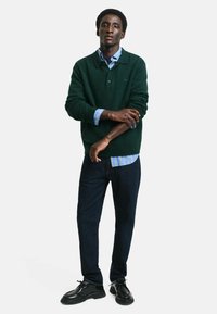 Grüner Strickpullover über einem blau gestreiften Hemd, dunkle Jeans und schwarze Schuhe. Entspannter Schnitt mit gerippten Bündchen und Saum. Kontrastierende Muster sichtbar.