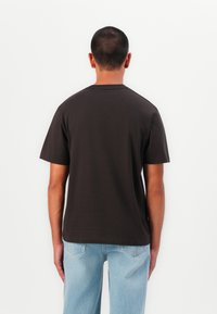 Dark brown short-sleeve t-shirt with a relaxed fit, made of cotton. Features a crew neck and smooth texture. Worn with light blue jeans.
