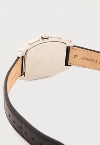 Πίσω μέρος ορθογώνιας ασημένιας θήκης ρολογιού με κορόνα και κουμπιά, λουράκι από ταμπά δέρμα με μαύρο διάτρητο εξωτερικό στρώμα.