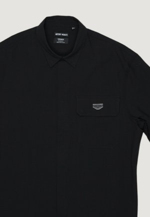 Camicetta da uomo nera con collo a punta, taschino sul petto con una piccola targhetta rettangolare in metallo e etichetta in tessuto all'interno del collo.