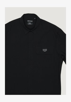 Chemise noire pour hommes à boutons avec col pointu, poche poitrine ornée d'une petite plaque métallique rectangulaire, et étiquette en tissu à l'intérieur du col.