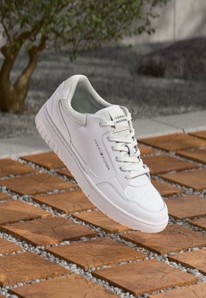 Biały tenis Tommy Hilfiger z sznurowadłami, umieszczony na drewnianej powierzchni z kafelkami z małymi kamieniami w szczelinach, obok małego drzewa.