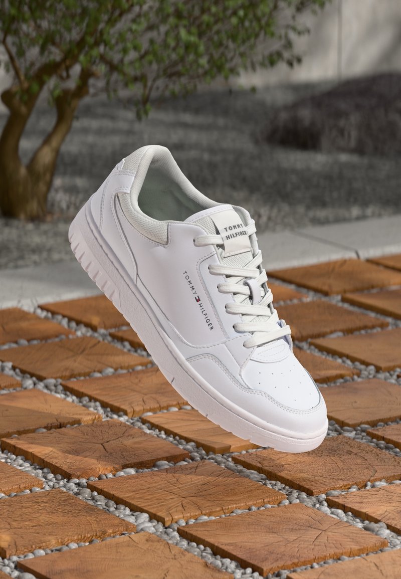 Baskets blanches Tommy Hilfiger avec lacets, placées sur une surface carrelée en bois avec de petits cailloux dans les interstices, à côté d'un petit arbre.