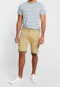 Beige cargoshorts met zijzakken en een ronde patch. Gepaard met een grijs-wit gestreept t-shirt en witte sneakers.