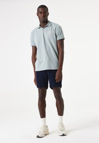 Camisa polo de color azul claro con un pequeño logotipo, combinada con pantalones cortos de textura azul marino. El modelo lleva calcetines blancos y zapatillas sport de color neutro claro.