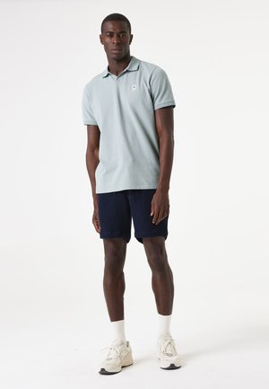 Camisa polo de color azul claro con un pequeño logotipo, combinada con pantalones cortos de textura azul marino. El modelo lleva calcetines blancos y zapatillas sport de color neutro claro.