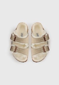 Beige sandaler med to justerbare stropper med spenne, et mykt pelsfôr og en teksturert såle for godt grep.
