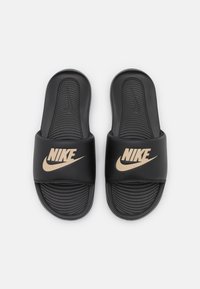 Crne Nike natikače s teksturiranom podlogom za stopala i širokom trakom s zlatnim logotipom. Glatki, gumeni materijal, dizajn otvorenog prsta i zakrivljen oblik.