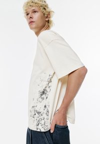 Tricou cu mânecă scurtă de culoare crem, din bumbac, cu un model texturat de stropi negri pe partea stângă și o croială lejeră.