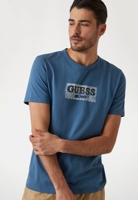 Modré bavlněné tričko s krátkým rukávem, se vzorovaným logem "GUESS" uprostřed. Výstřih je kulatý a střih je standardní.